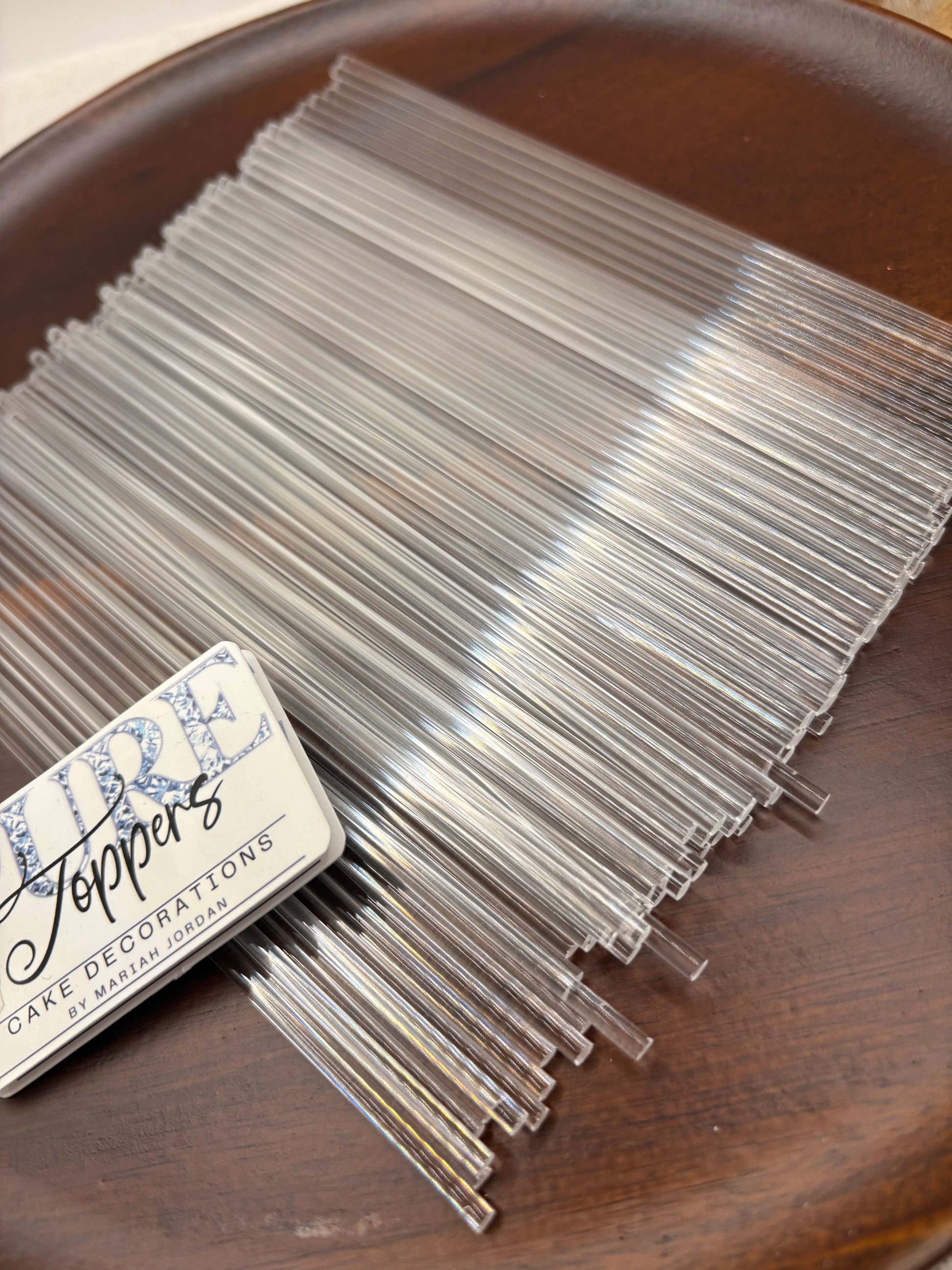 A bundle of clear glass cake toppers, including picks and flat decorative pieces, arranged on a dark brown wooden plate next to a metal tin labeled 'Pure Peppers Cake Decorations'.