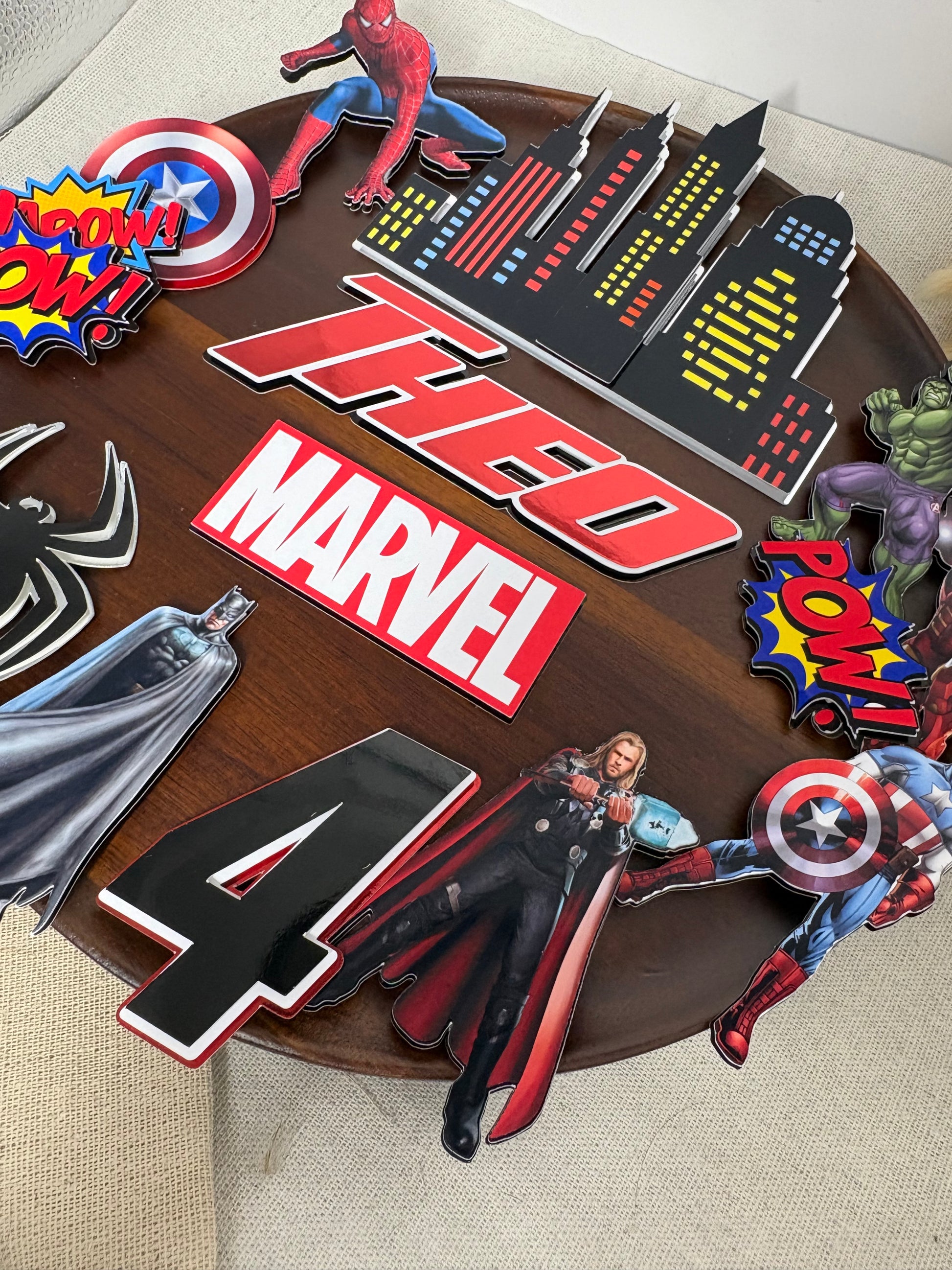 A round wooden birthday cake decorated with Marvel-themed cutout decorations, including Spider-Man, Captain America, Thor, Hulk, Black Widow, and a large 'THED MARVEL' sign, with a black number 4 on the side.
