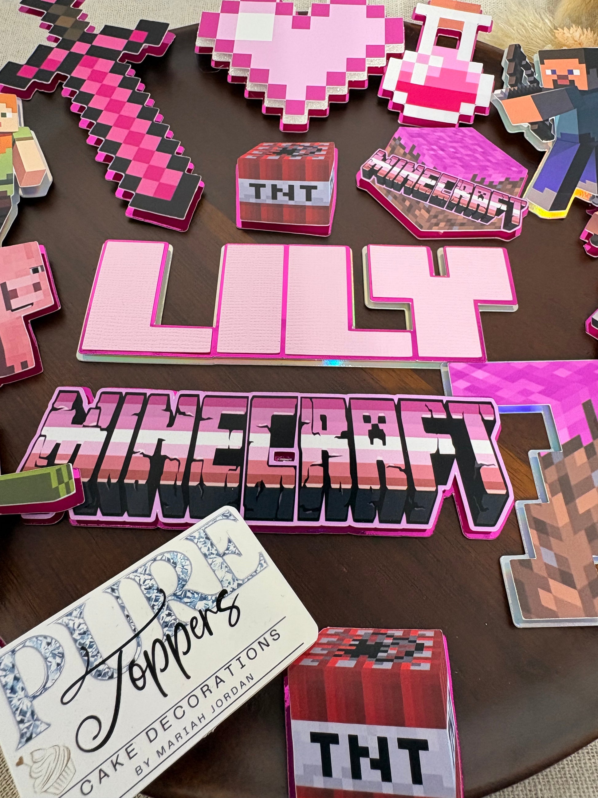 A collection of handmade Minecraft-themed decorative cutouts and 3D letters arranged on a dark brown tabletop. The designs feature pink, black, and white pixelated block graphics, including the word 'MINECRAFT', a sword, a heart, TNT boxes, and a decorative card.