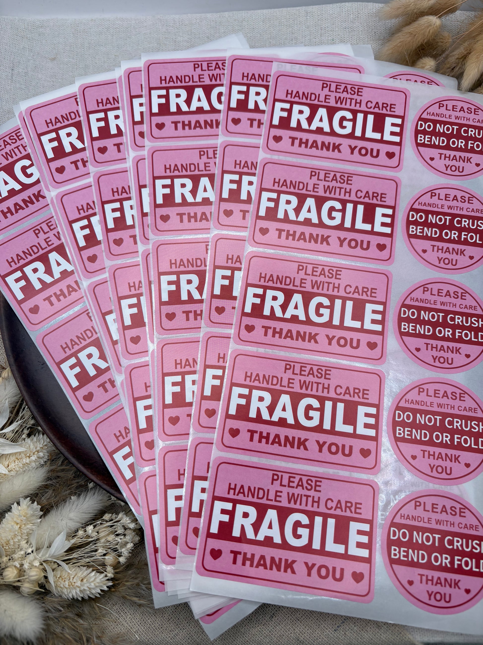 Pink and red 'FRAGILE' handling stickers arranged in overlapping sheets on a neutral fabric background with dried floral props.
