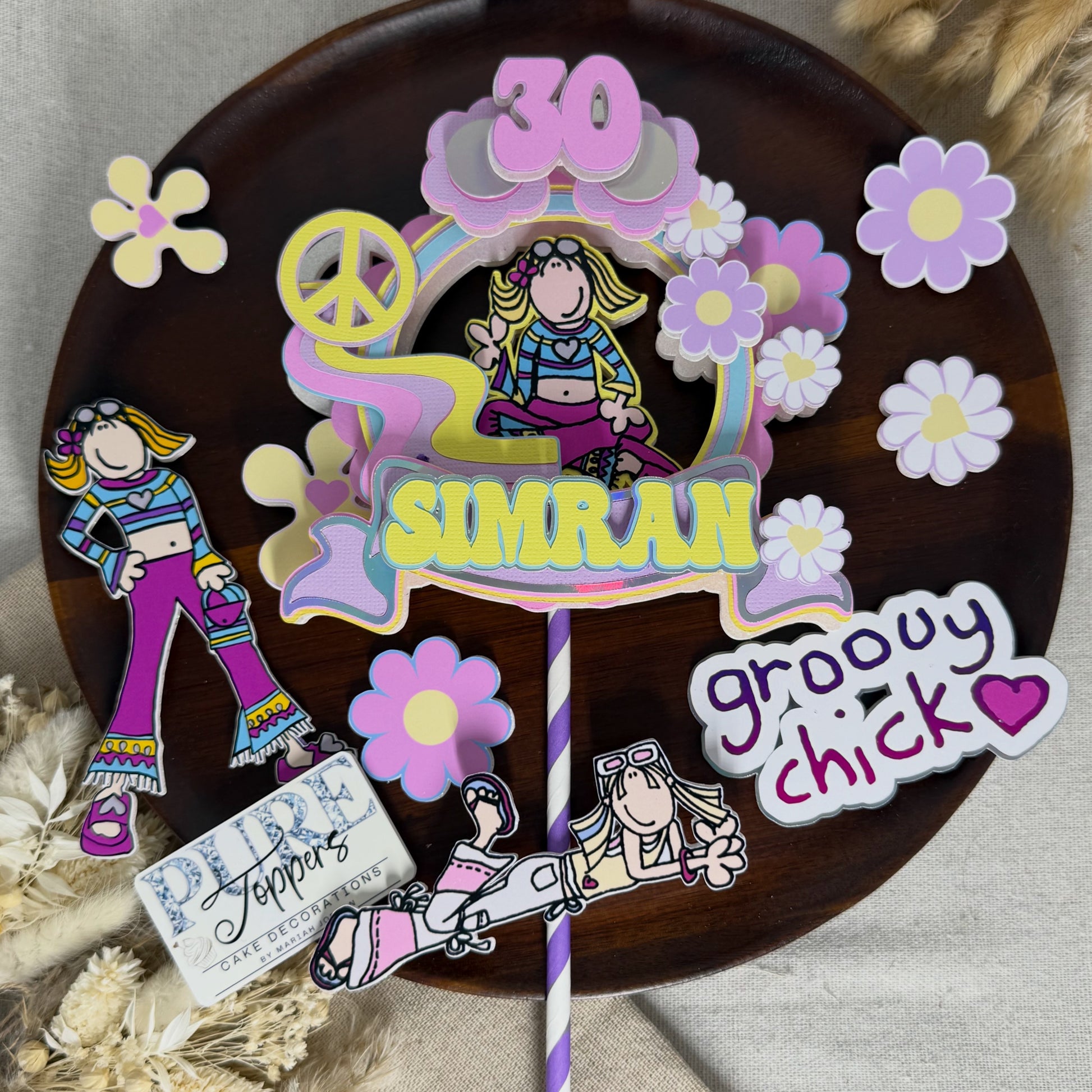 A custom paper cutout birthday cake topper featuring a retro 1970s/80s theme. The design includes a cartoon girl in colorful striped clothing, a rainbow arch, a peace sign, a pink number 30, the name 'SIMRAN' in yellow, and a 'groovy chick' sticker.
