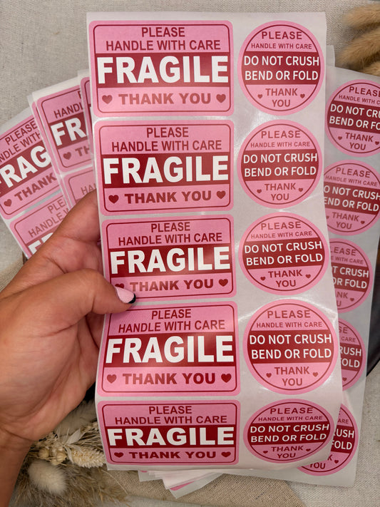A hand holds a sheet of pink and white shipping stickers with 'FRAGILE' text, alongside other sheets showing additional designs on a neutral background.