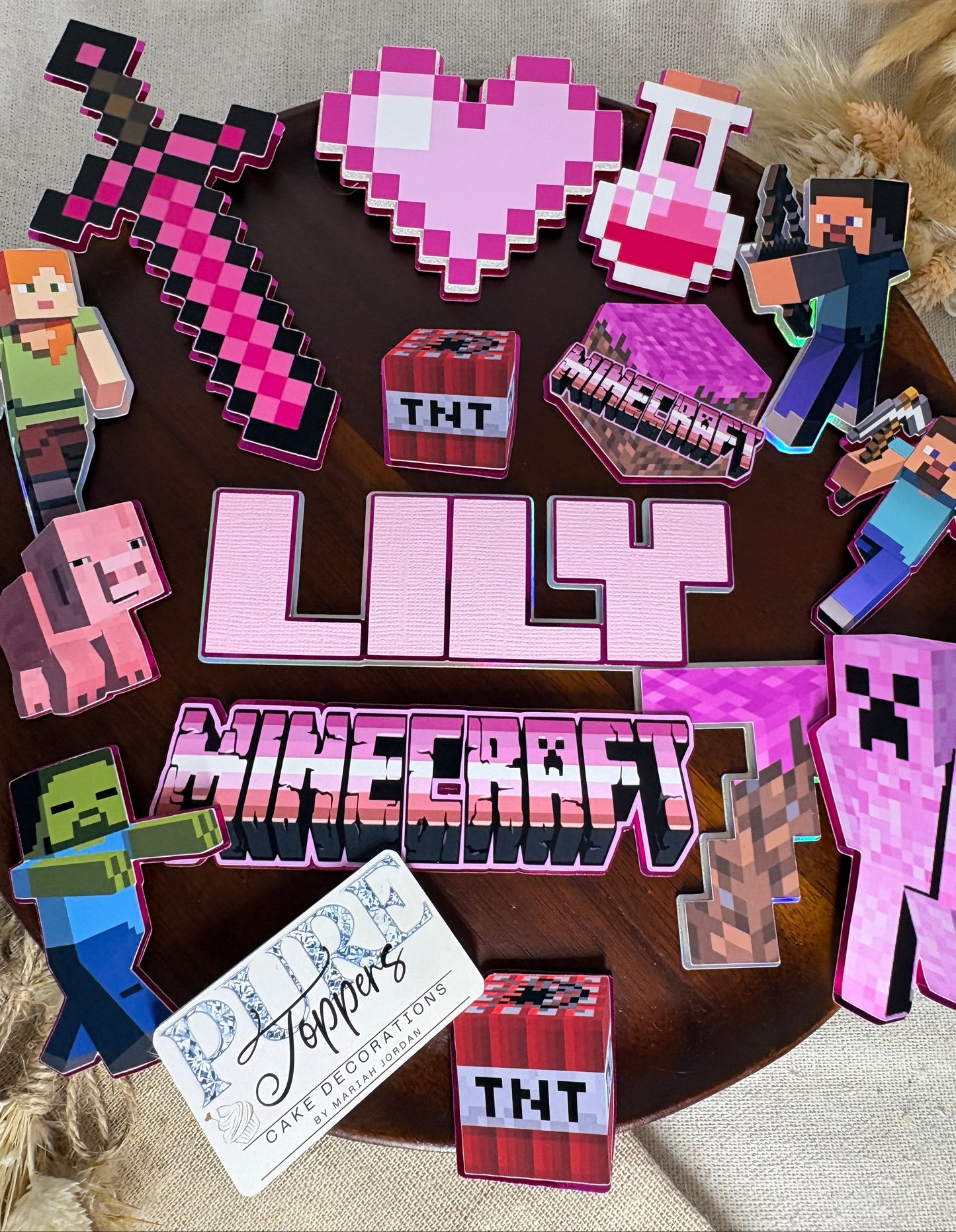 A collection of custom Minecraft-themed paper cutouts and stickers displayed on a dark round table. The set includes a large pink 'LILY' nameplate, a vertical 'MINECRAFT' sign, a pink pixelated heart, a sword, a potion bottle, a Creeper, a Zombie, a Pig, a Steve character, TNT boxes, and a custom card reading 'Sedde'.