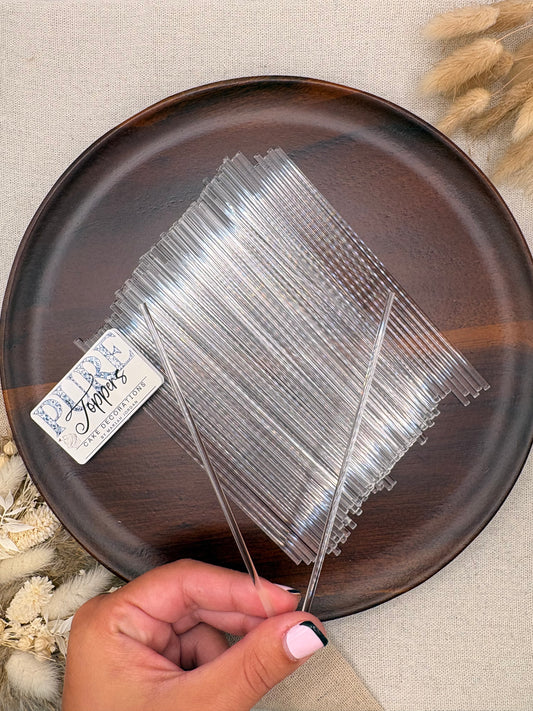 A hand holds a clear plastic strainer mesh or filter mat over a dark brown ceramic plate, with a small white brand tag visible. The scene is styled on a beige fabric background with dried flowers.