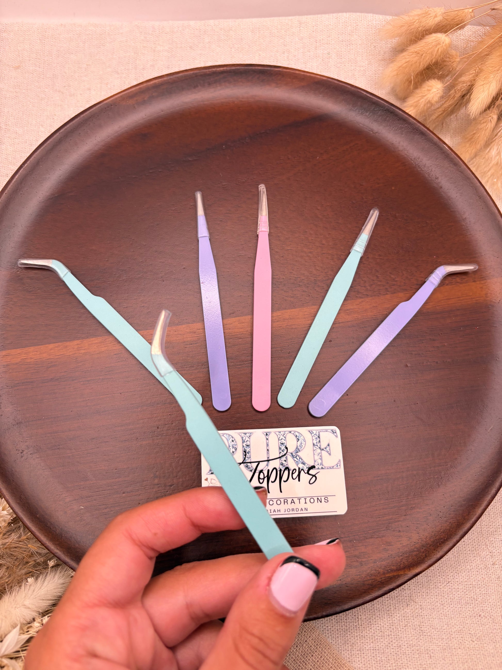 A hand with pink nail polish holds a light blue curved metal tweezer over a round dark brown wooden tray. Four more metal tweezer tips in pastel purple, pink, and mint green are arranged on the tray next to a small white card.