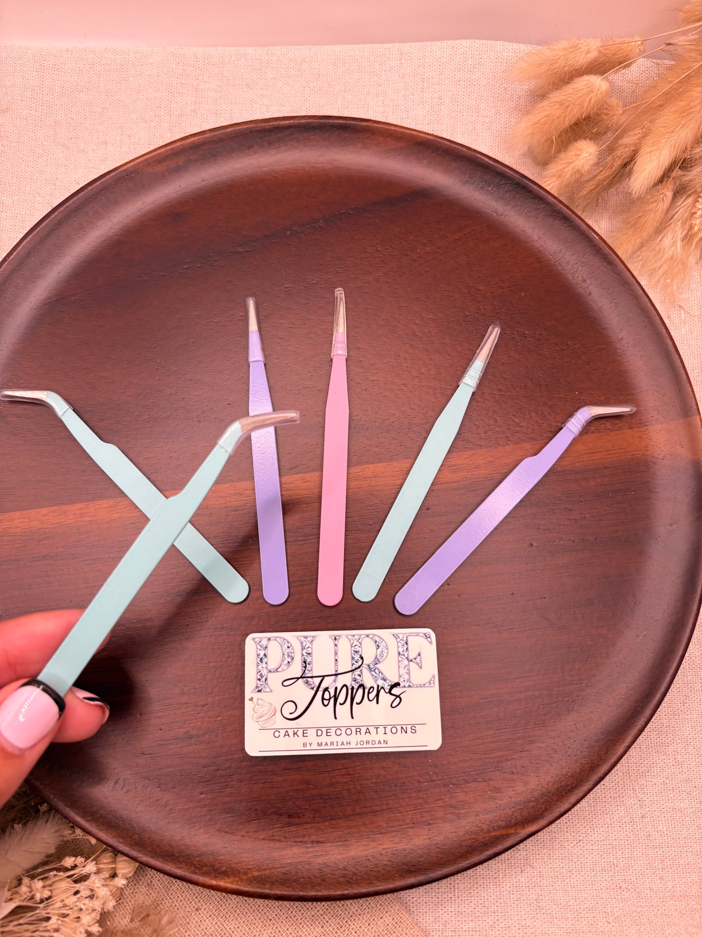 Four pastel-colored plastic cake decoration tweezers arranged on a round dark brown wooden plate, with a hand holding one mint green tweezzer in the foreground. A small white card with the brand name 'Soppy's Cake Decorations' is placed on the plate.
