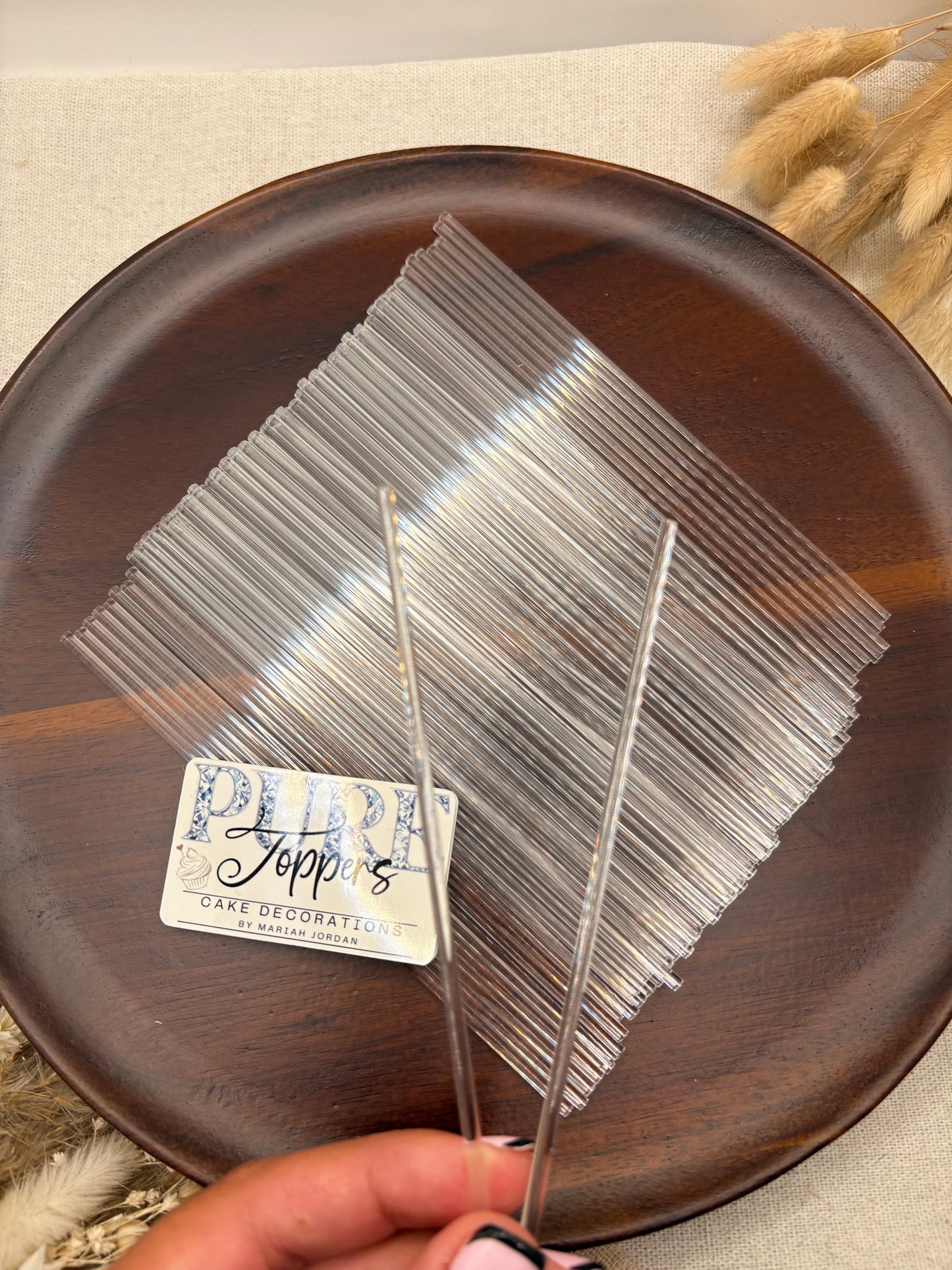 A hand holds two clear plastic cake toppers over a stack of similar clear plastic pieces on a round dark wooden plate. A small card reading 'Pete Toppings' is visible near the items.