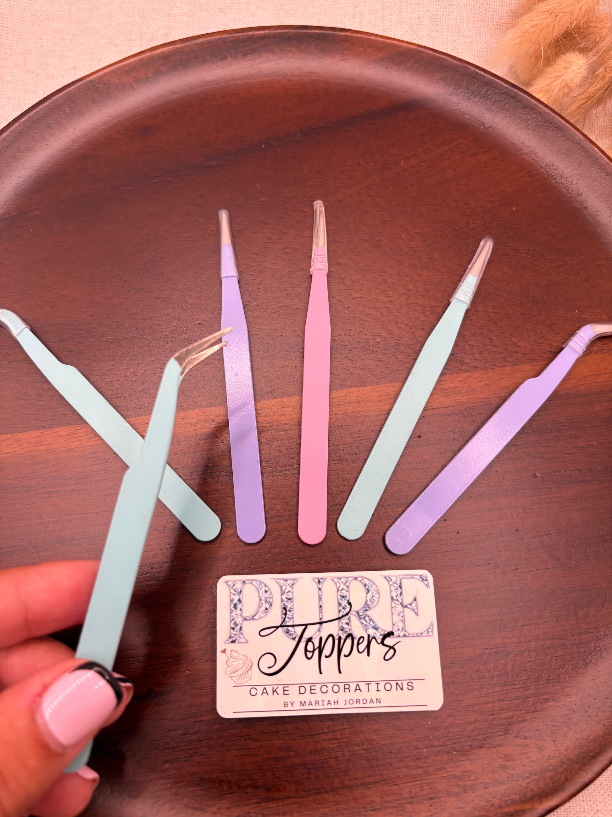 A hand with pink manicured nails holds a pair of pastel blue tweezers over a dark brown wooden tray, alongside other pastel-colored tweezers and a small card reading 'Joppers Care Decorations'.