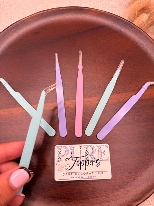 A hand with pink manicured nails holds a pair of pastel blue tweezers over a dark brown wooden tray, alongside other pastel-colored tweezers and a small card reading 'Joppers Care Decorations'.