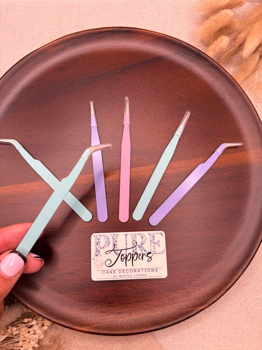 Five pastel-colored plastic tweezers arranged on a round dark brown wooden plate, with a small card reading 'Leppins Cake Decorations' and a hand holding one light blue tweezzer at the bottom right.
