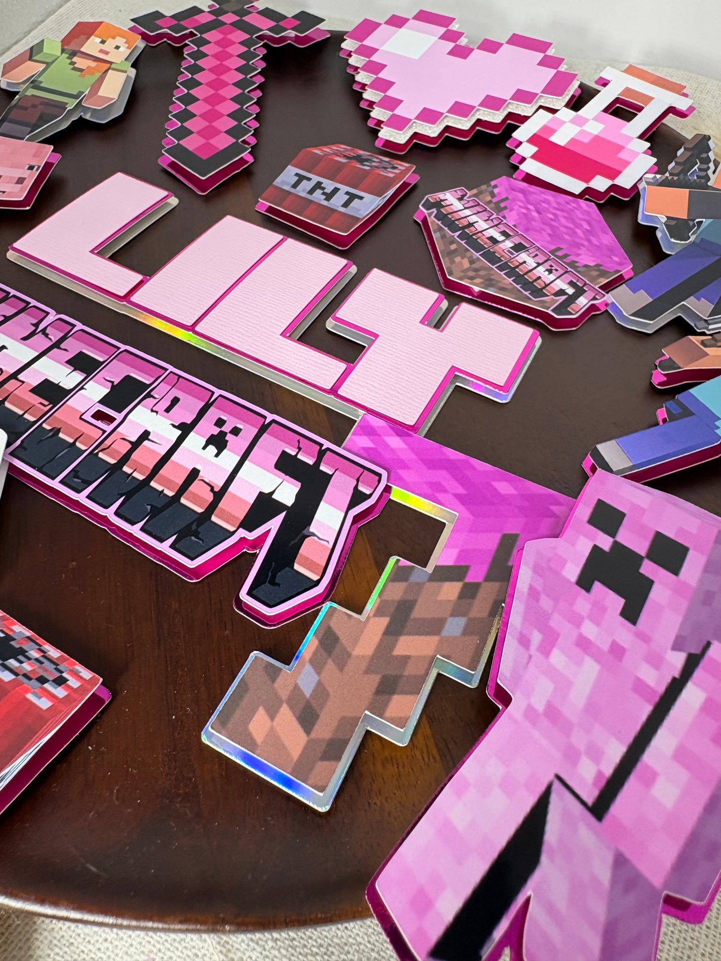 A collection of pink and purple Minecraft-themed die-cut stickers and cutouts arranged on a dark brown wooden surface, including a pixelated sword, TNT, a creeper, a player character, and stylized text.