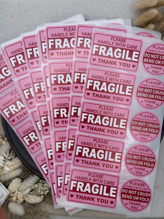 A stack of pink and white 'Fragile' shipping labels with red text, arranged on a neutral fabric background with decorative dried flowers.