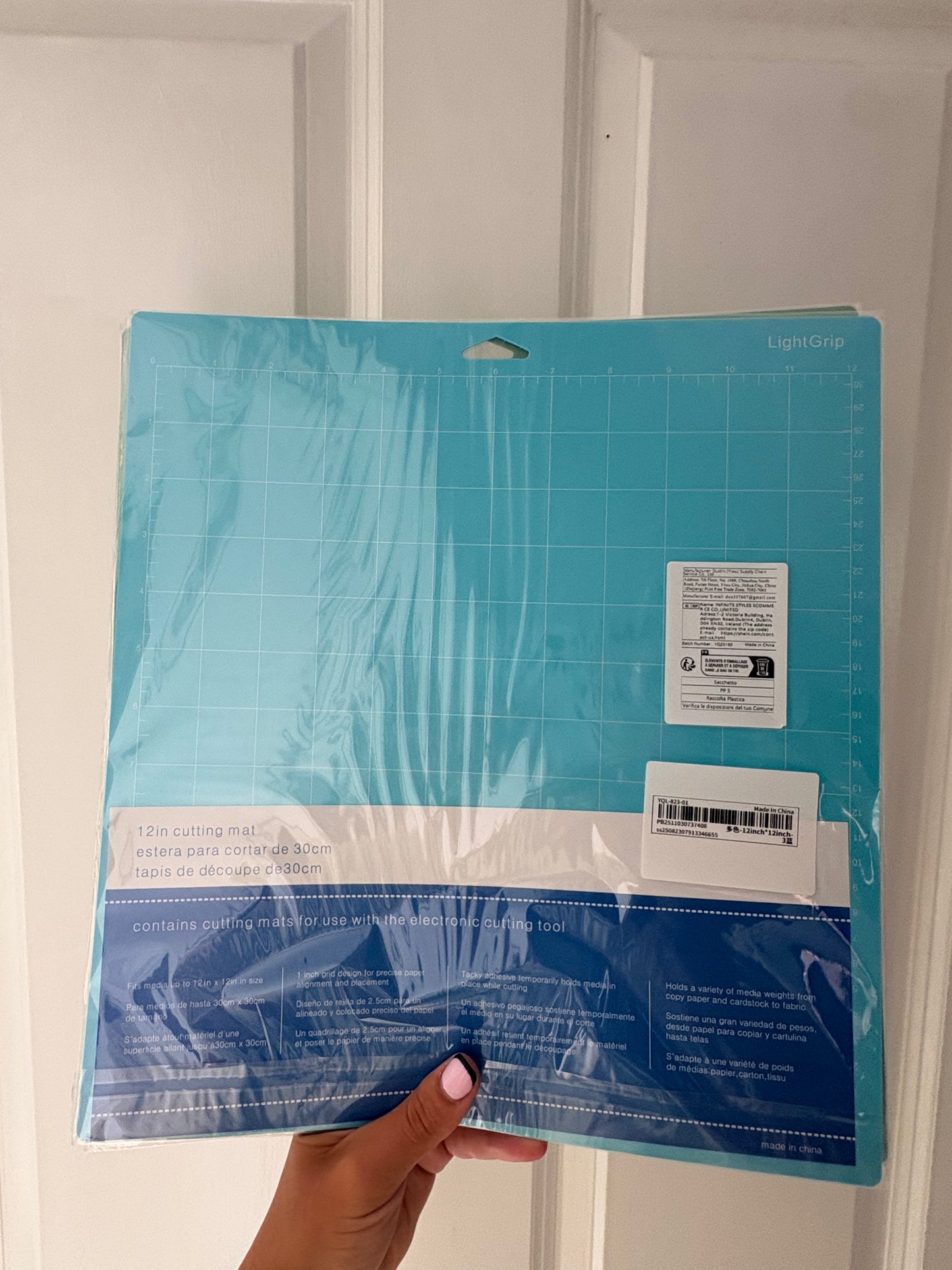 A 12x12 inch Light Grip cutting mat in teal blue, still in its retail plastic packaging, held by a hand against a white paneled door background.