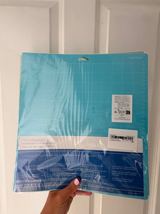 A 12x12 inch Light Grip cutting mat in teal blue, still in its retail plastic packaging, held by a hand against a white paneled door background.