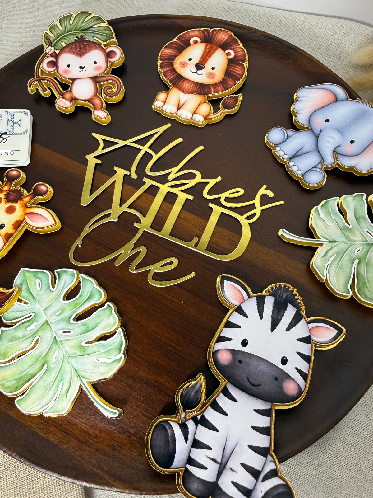 A set of colorful safari-themed cookie cutters displayed on a dark wooden round board. The cutters feature cartoon animals including a zebra, elephant, lion, monkey, giraffe, and a zebra, along with green tropical leaves and a gold script lettering piece that reads 'Albie's Wild One'.