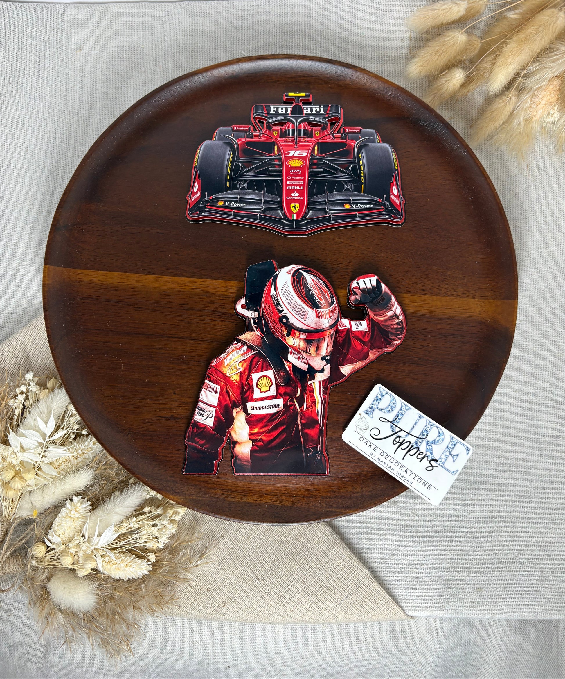 A round dark brown wooden serving tray with two Ferrari-themed decals, one of a red racing car and another of a driver in a red racing suit, displayed on a beige fabric background with dried flowers.