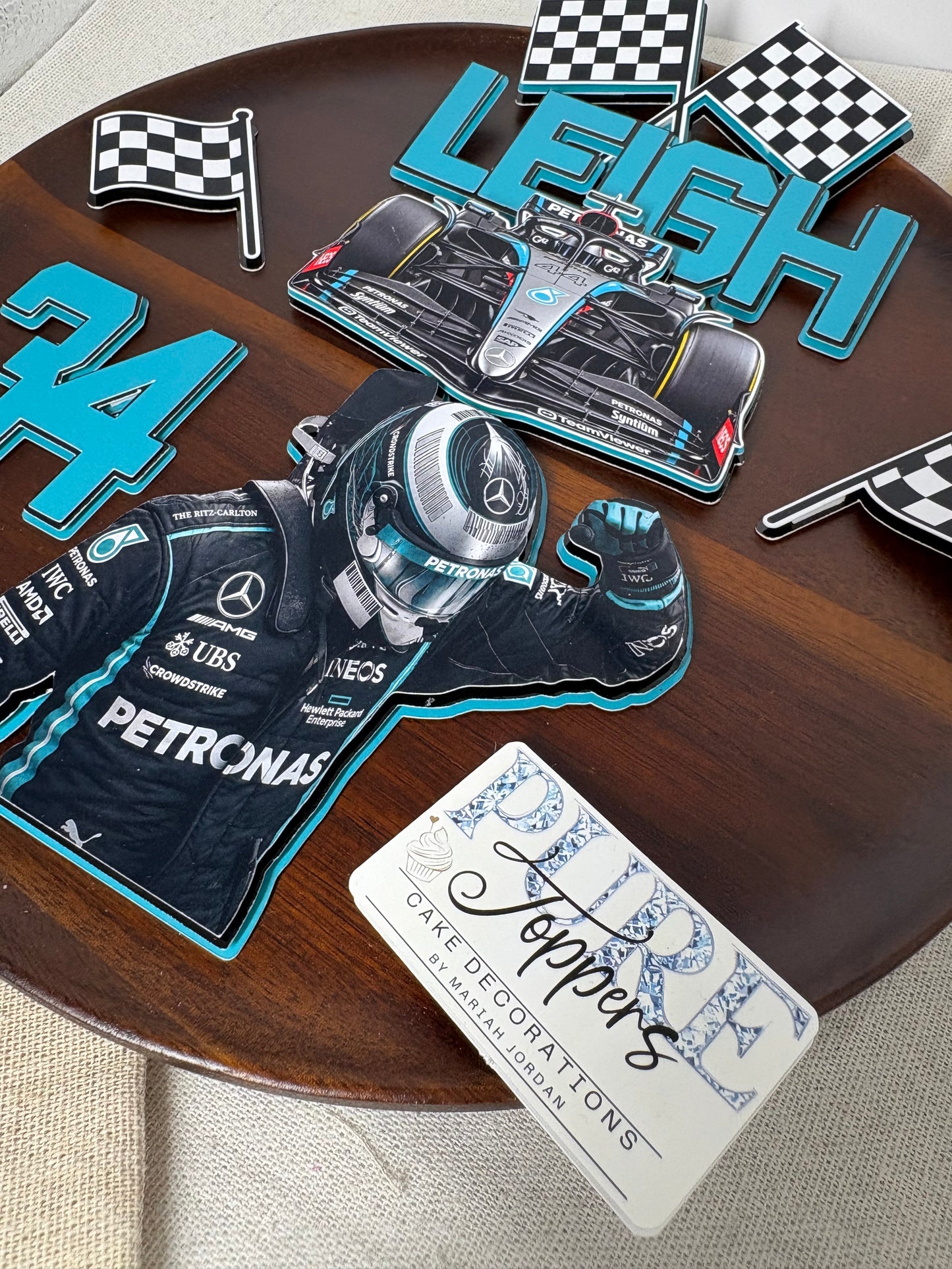 A set of custom racing-themed paper cutout toppers arranged on a round wooden table. The set includes a large 'LEIGH' topper with a Mercedes F1 car graphic, a large '24' topper, a cutout of Lewis Hamilton in a Petronas racing suit and helmet, several checkered flags, and a white card reading 'RIDE Toppers' with 'ONE DECORATIONS'.