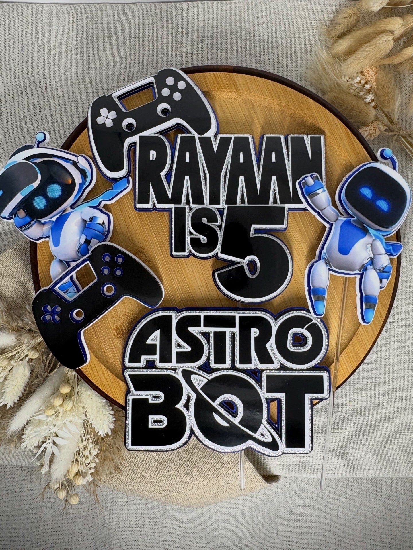 A personalized 5th birthday topper sign reading 'RAYAAN IS 5 ASTRO BOT' in bold black letters with white outlines, decorated with two blue and white robot characters and black video game controllers, displayed on a round wooden board with dried floral decorations.